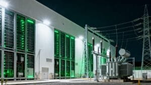 A modern data center building with illuminated green server racks stands next to an electrical transformer and high-voltage power lines at night.
