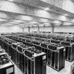 Large industrial server room with multiple rows of computer servers and extensive overhead piping for cooling, in a concrete-floored facility.