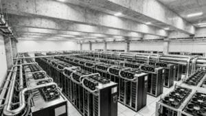 Large industrial server room with multiple rows of computer servers and extensive overhead piping for cooling, in a concrete-floored facility.