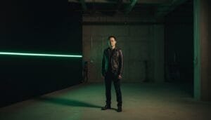 A person wearing a black leather jacket and dark clothing stands in a dimly lit, industrial space with a green horizontal light strip on the wall.