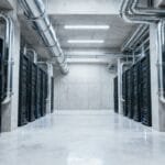 A server room with rows of black server racks, exposed pipes on the ceiling, and a clean, reflective concrete floor.