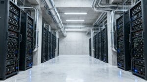 A server room with rows of black server racks, exposed pipes on the ceiling, and a clean, reflective concrete floor.