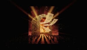 A crowd faces a large, fiery structure with burning currency symbols as papers with dollar, euro, and yen signs fly out, lit dramatically by intense beams of light.